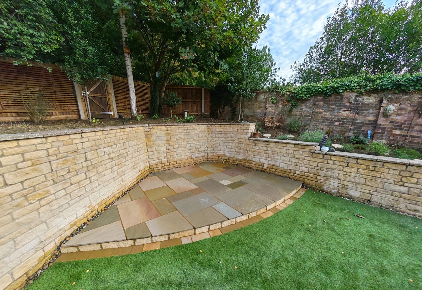 Multi-tone Indian sandstone patio with a curved raised edge in the corner of a walled garden in Weldon Multi-tone Indian sandstone patio with a curved raised edge in the corner of a walled garden in Weldon